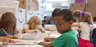 Enfants dans un local de classe assis à leur bureau travaillant concentrés, avec un petit garçon en t-shirt vert qui porte des lunettes en premier plan