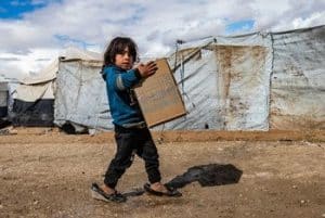 Ett barn som bär på en låda i en miljö som liknar ett flyktingläger
