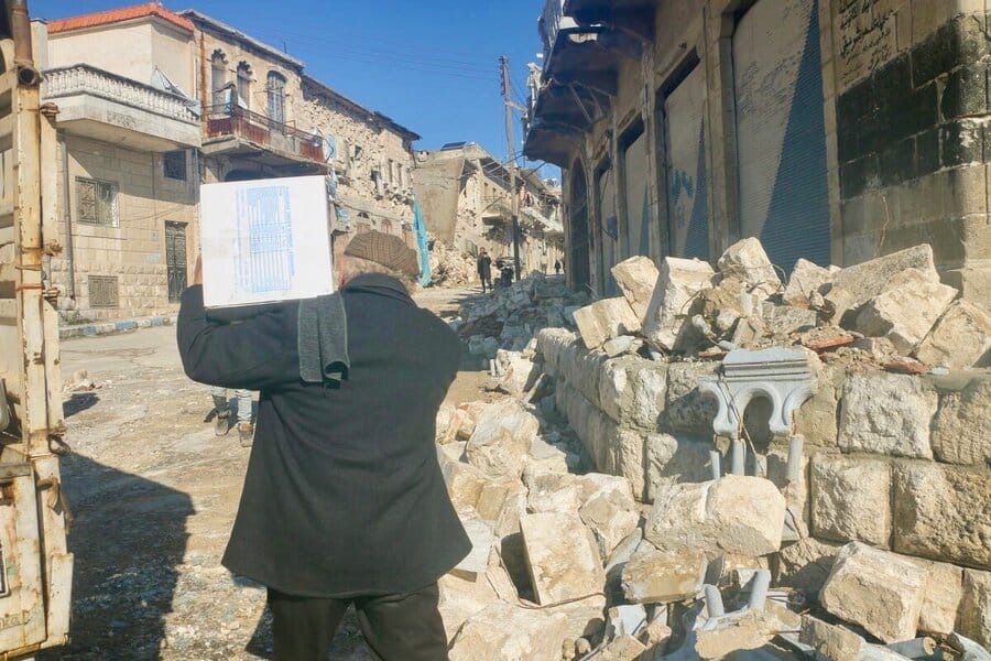 En man som bär på ett paket med ryggen mot kameran. I bakgrunden förödelse och förstörda byggnader.