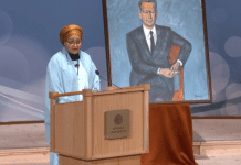 En kvinna bakom ett podium som håller ett tal. Bredvid en tavla av en man.