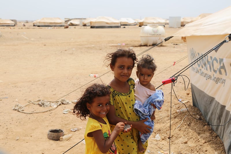 Tre barn som står framför ett flyktingläger på en öppen sandig plats .