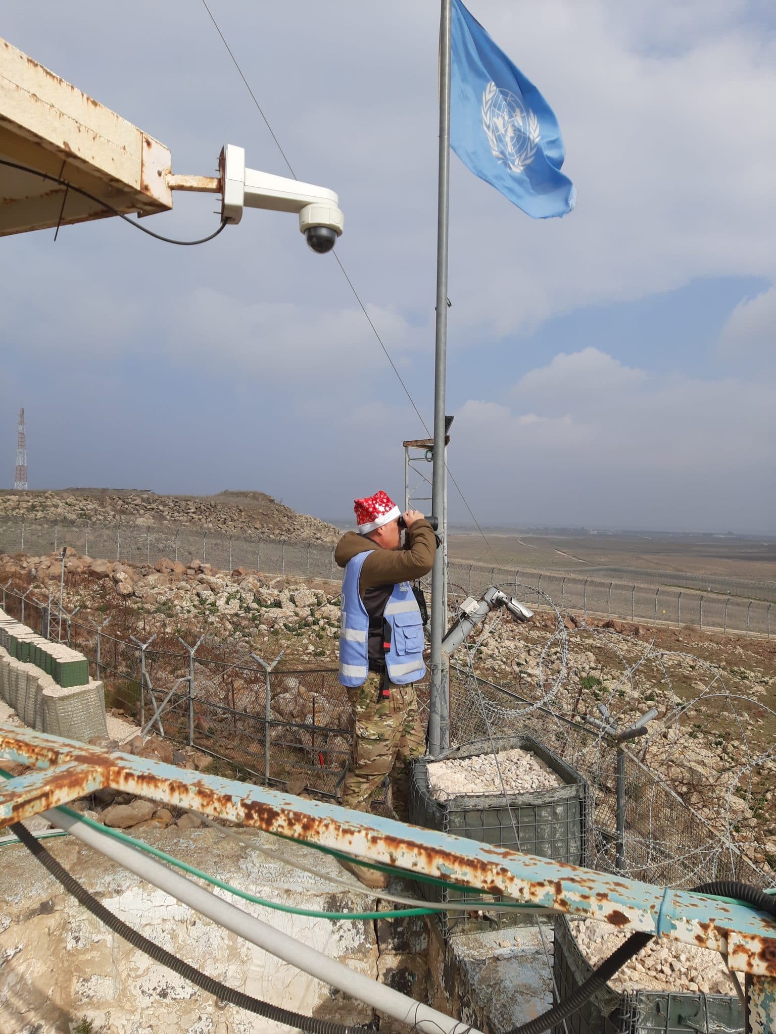 En man i uniform som står vid FNs flagga och tittar i en kikare